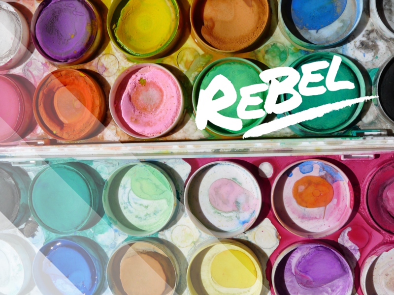 Watercolor paints in a palette with the word 'REBEL' scribbled over, symbolizing creativity and unconventional thinking.