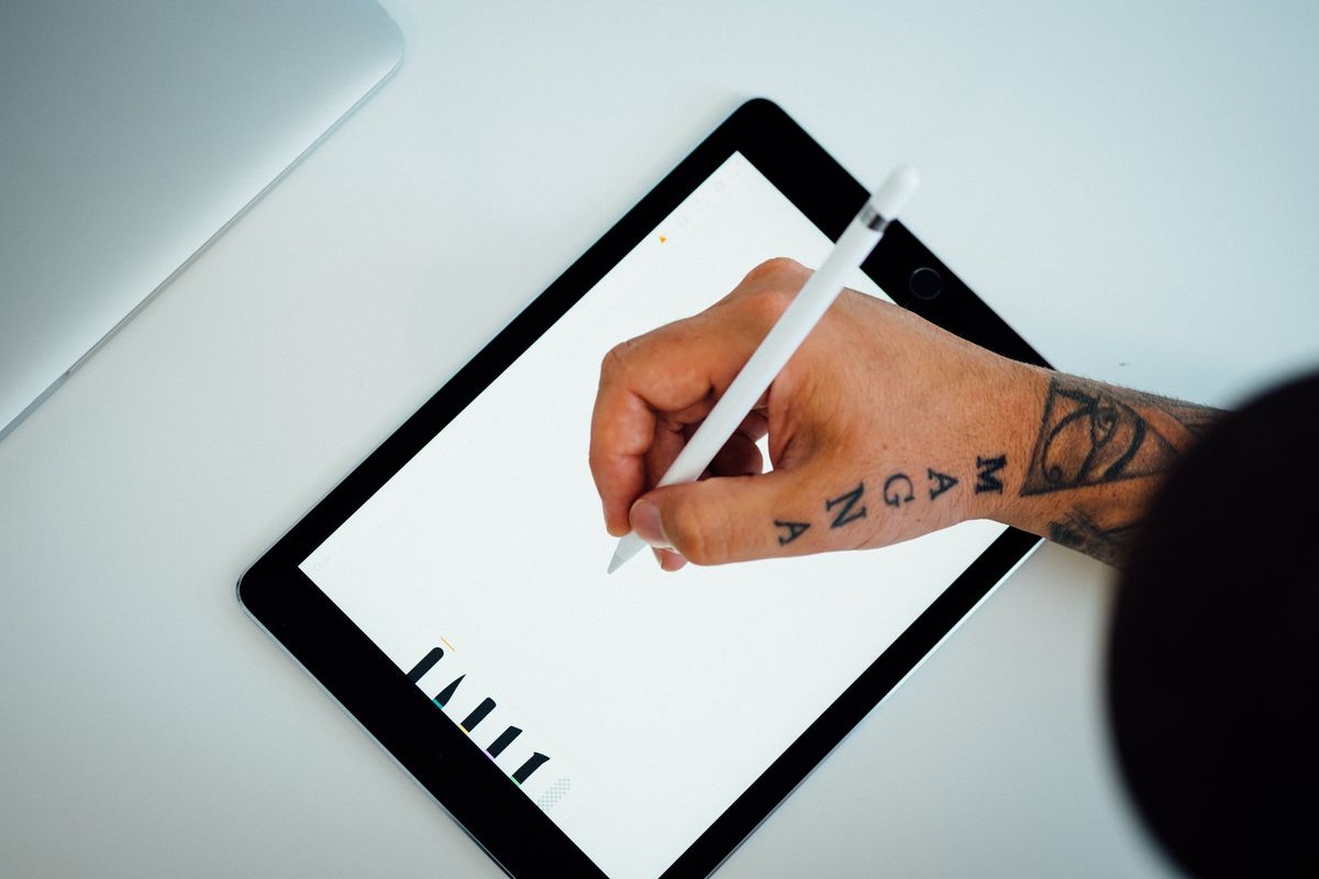 Close-up of a tattooed hand writing on a digital tablet, representing the adaptability of using an iPad Pro for work.